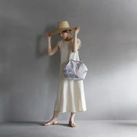 Woman in a white dress and straw hat holding a patterned bag against a gray background